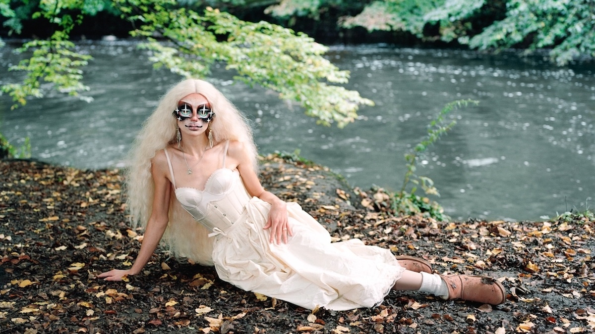 Sgaire Wood reclining at a pebbly riverbed, she is wearing a white corseted dress, sparkling eye makeup, and long curls.