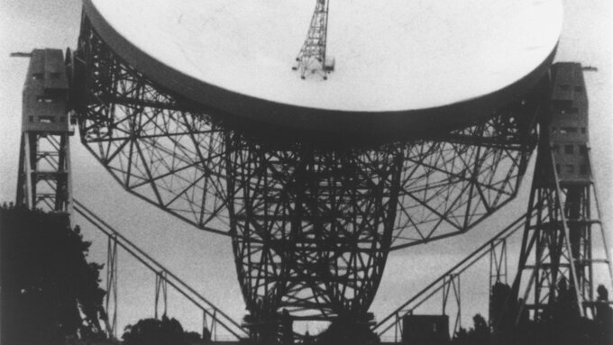 A black and white film still of a huge radio telescope. The white dish is angled upwards.