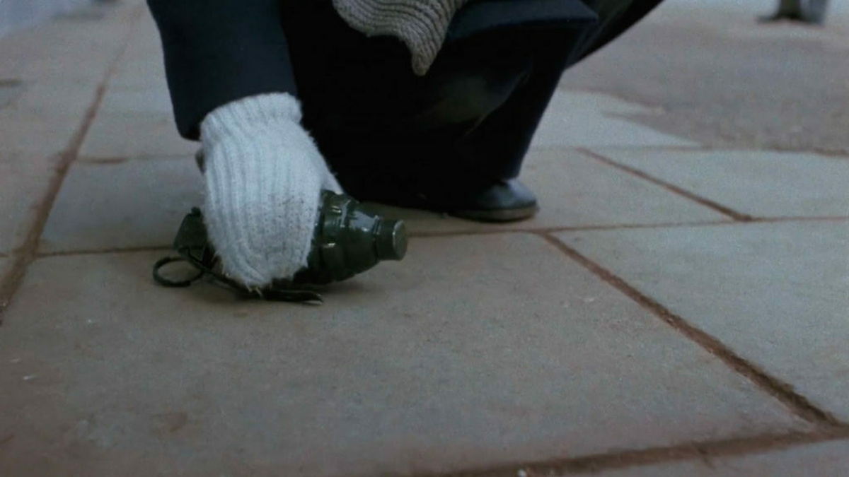 A hand in woolen glove places a hand grenade on a pavement.