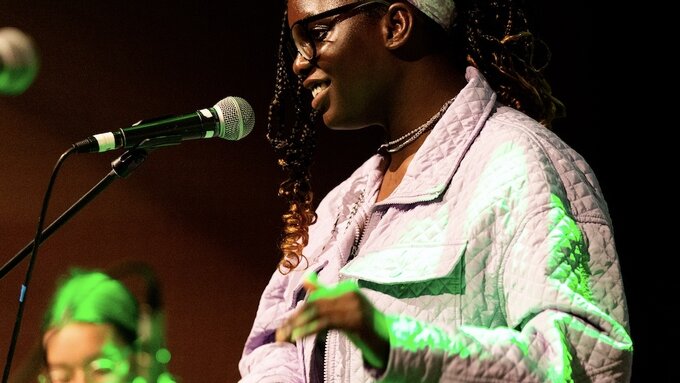 A smiling girl dressed in purple sings into microphone, her mentor is in the background playing keyboard.