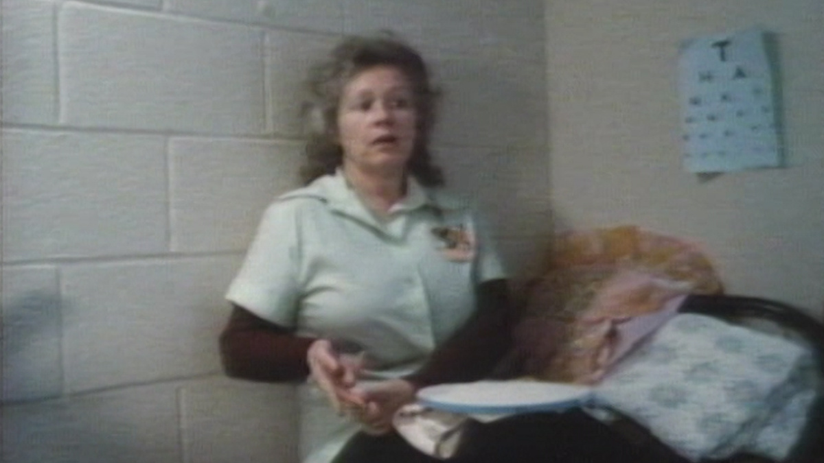 A woman sitting on a bed with her back to the wall.  The image is grainy and slightly out of focus.