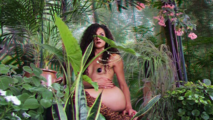 Performer Mavi Veloso sat mostly nude in a forested area, it is full of lush greens and tropical plants.