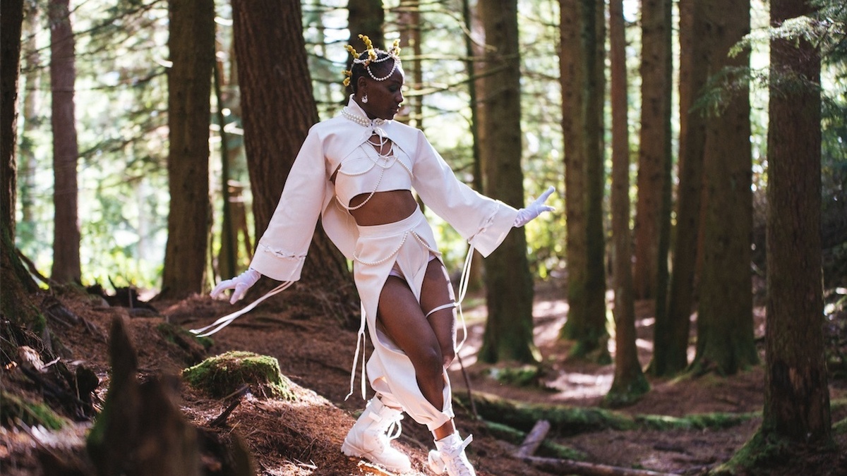 A black femme person wearing a stylish all white outfit, reminiscent of a cropped suit, walking through a forest.