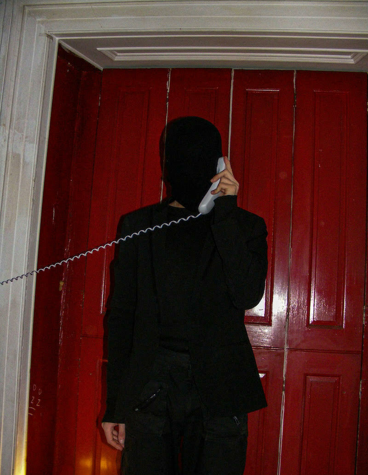 HUSS Stood in front of a red door, wearing an all black body suit holding a white corded phone to his ear.