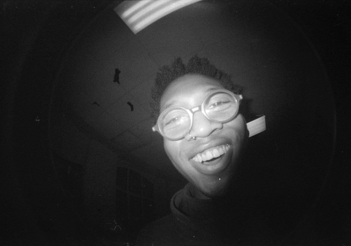 A dark photo of Chizu Nnamdi, a black person wearing round thick rimmed glasses. They smile down at the camera.