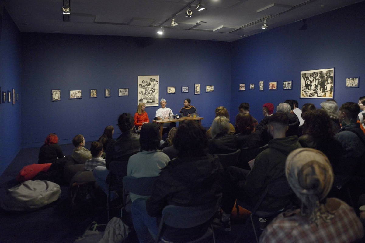 Three people sit at a table in a blue gallery, lots of people sit on chairs, listening to them speak.