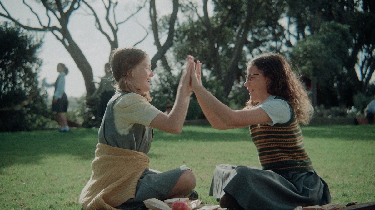 Two young girls in school uniform, sit on the grass and face each-other raising their hands to touch