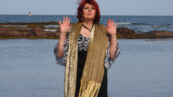 A person stands wearing a scarf, facing the camera with their hands up. There is a coastline and sea in the background.