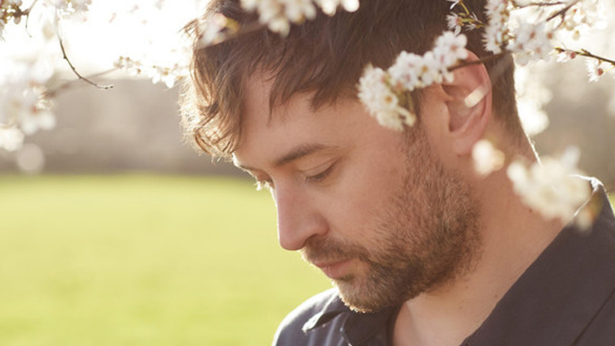 A man with a beard and brown hair looking down, his face is partially obscured by branches and tree blossom