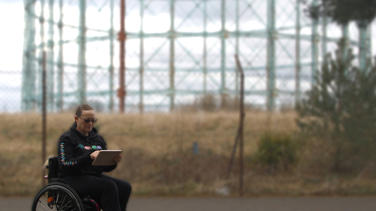 Clare Johnston in a wheelchair composing on an iPad, in front of a chainlink fence & blue gas container behind them.
