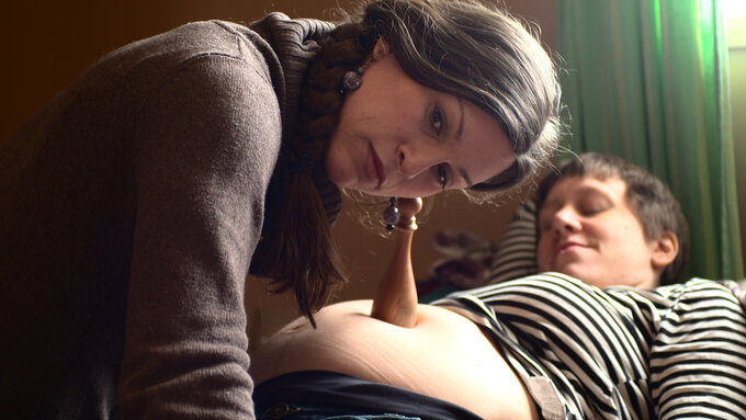 A still of a lesbian couple from the film. Jenna, a trans woman uses a tool to listen to her pregnant partners’ belly.