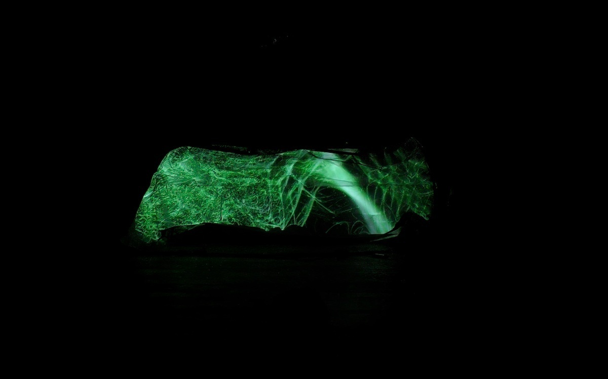 An operating table covered in fabric in a dark room with green projection mapping projected on to it.