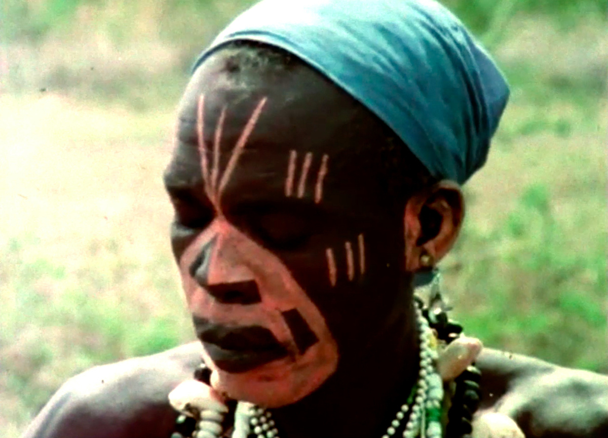 A black community leader undertaking spiritual practices. He is painted with orange markings & he wears bead jewellery.