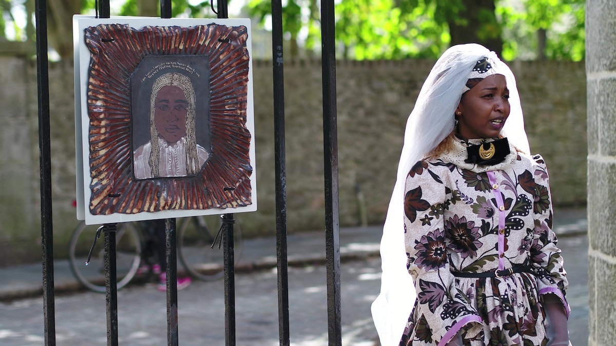 A black woman standing outdoors in a floral dress wearing a white vale next to an artwork tied to a fence.