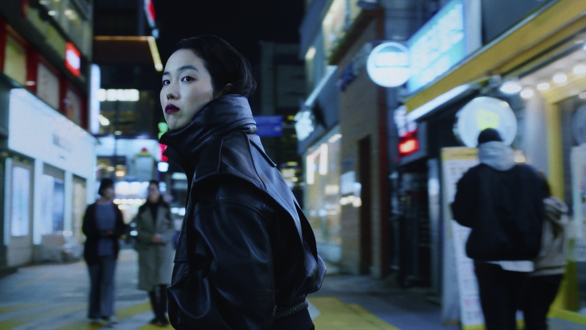 Film still: A woman with dark hair, wearing red lipstick and a leather coat, looks over her shoulder on a neon street.