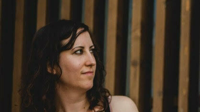 A portrait of poet J L Williams a white woman with long black wavy hair wearing a black vest