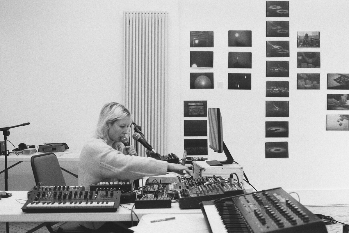 A black and white photo of a woman with blonde hair sat at a desk surrounded by mics, keyboards, and sound equipment.