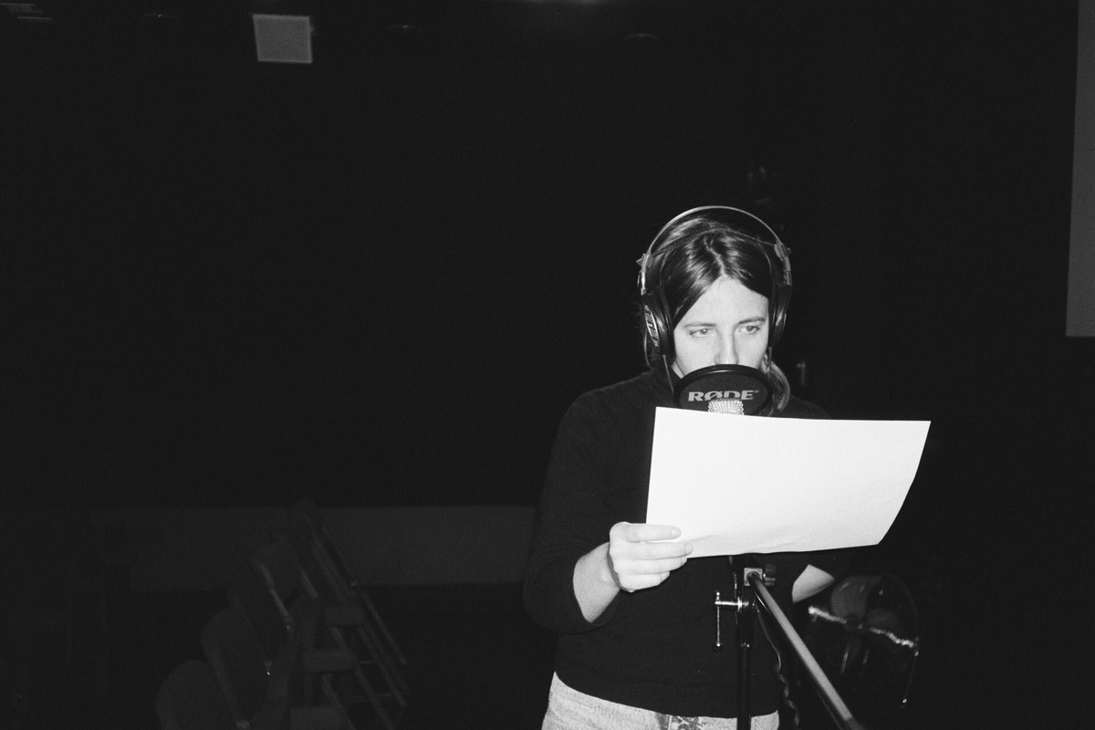 A dark black and white image with a dark haired person speaking into a microphone whilst reading from a sheet of paper.
