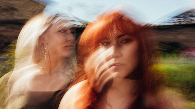 Two women standing outdoors. One women with red hair stares into the camera while the other looks over her shoulder.