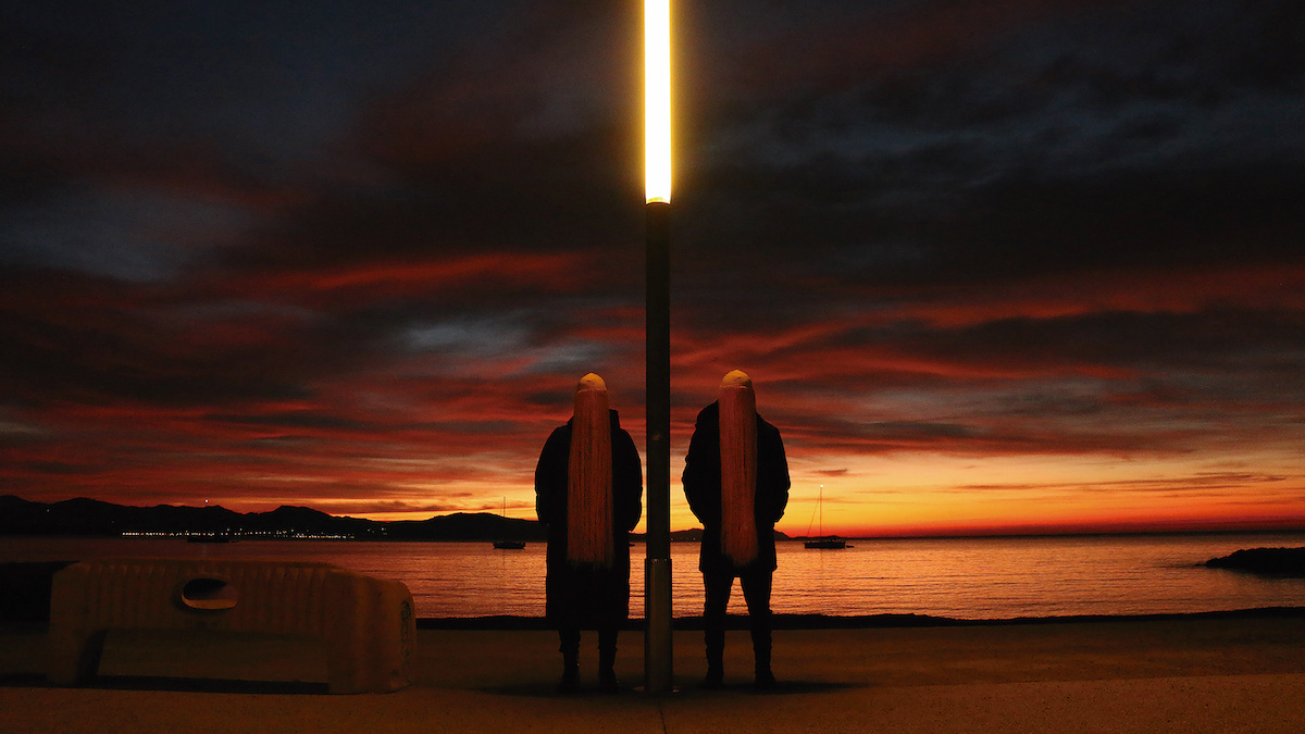 Two masked figures standing along either side of a street light against bay at sunset.