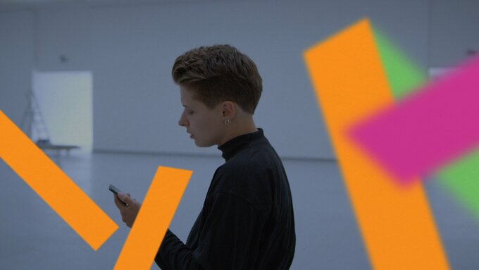 A person in a gallery space. They look down at their phone reading a text. The image is interrupted by colourful shapes