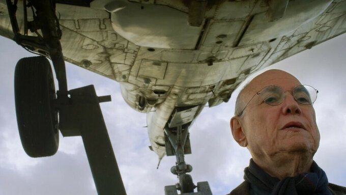 An elderly man's head below the hull of a fighter jet, a cloudy sky in the background.