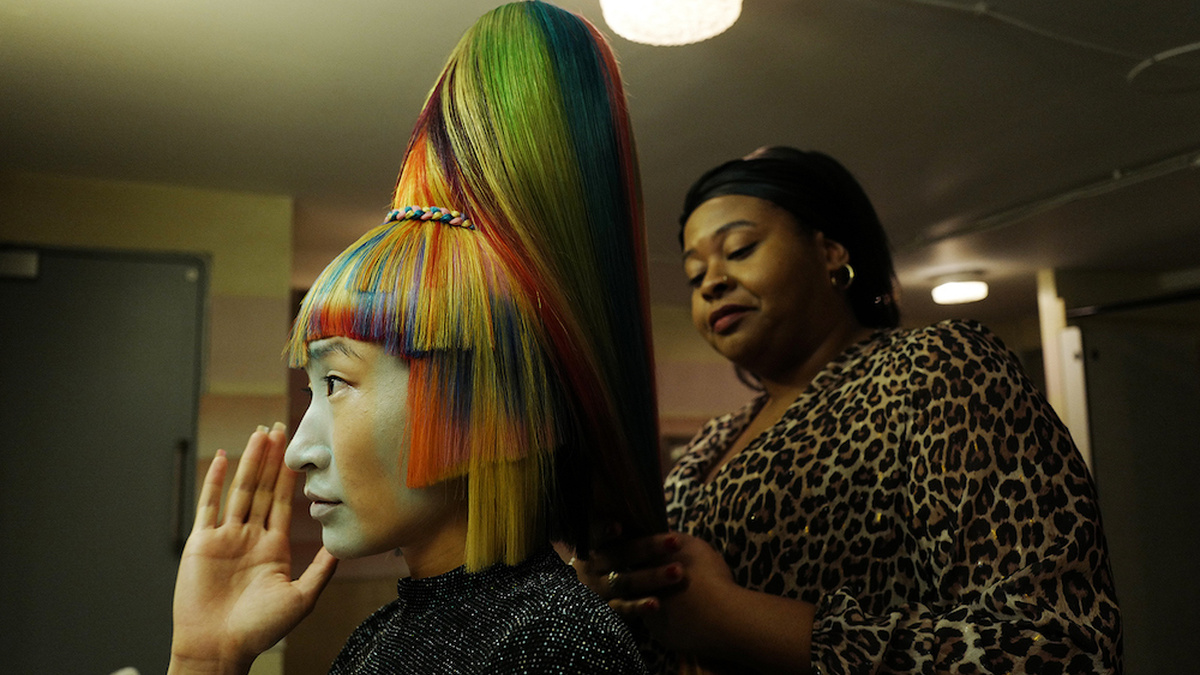 A hair model is having her hair styled in an elaborate way, with rainbow colours and a huge beehive by a hairdresser.