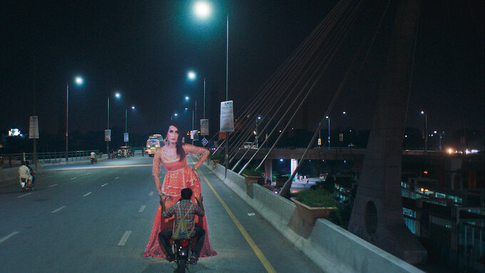 A night scene of a man holding the large cut out of a woman whilst riding a moped across a bridge.