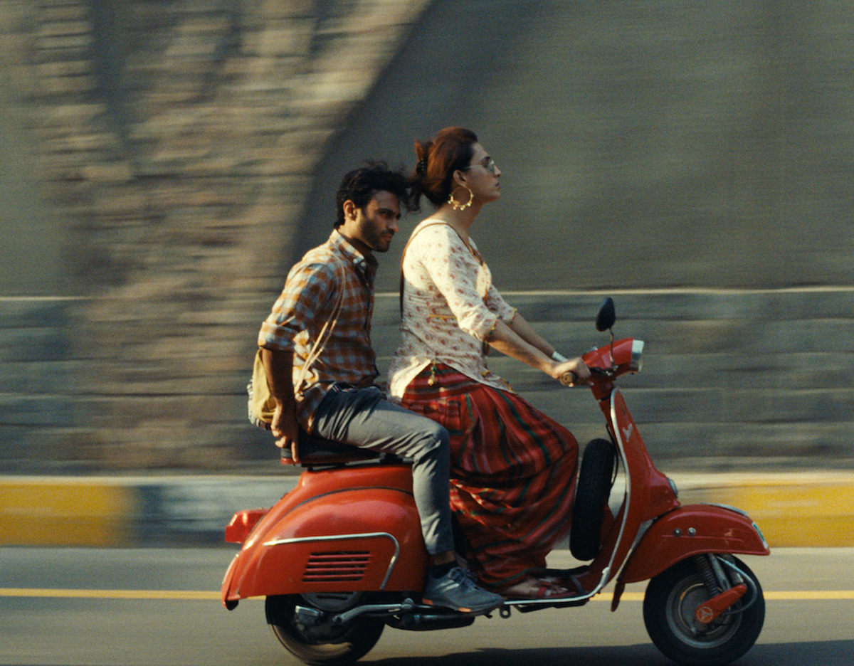 A woman driving a red moped down a street with a man riding on the back. The background is a blur.