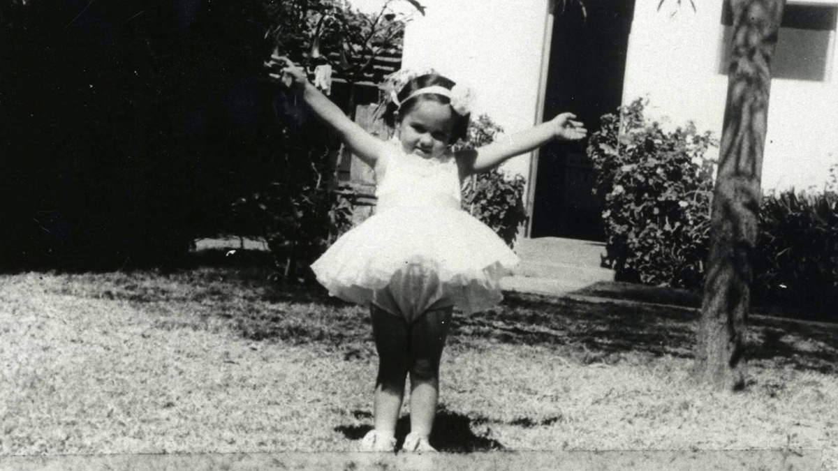 A black and white image of a young child in a ballet dress and extended arms posing for the camera in a back garden.