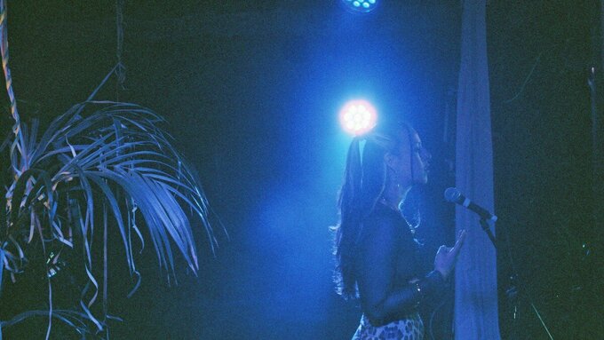 A girl with long, tied-back hair standing in front of a microphone in a blue lit room.