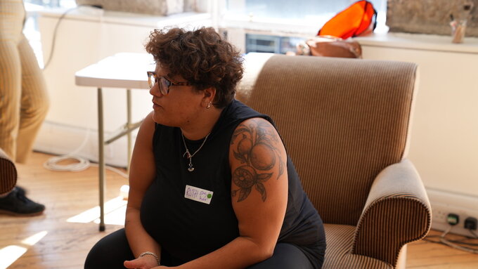 A woman with short hair and tattoos sitting on an armchair and leaning forward as if engaged in conversation.