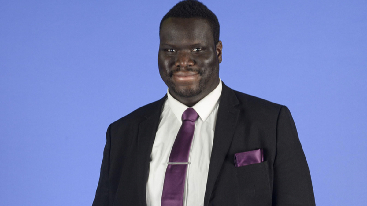 A black man in a smart suit with a purple tie standing and smiling looking towards the camera against a blue backdrop.