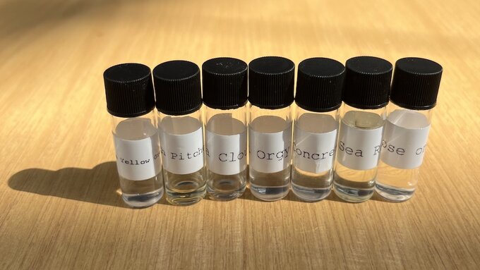 7 small clear bottles sit side by side on a pine desk top. Each bottle has a black screw top lid and a white label.