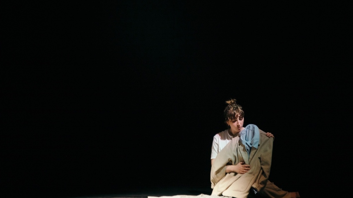 In a dark room a woman with a white t-shirt sits on the floor she is clutching a cream suit with a blue shirt underneath