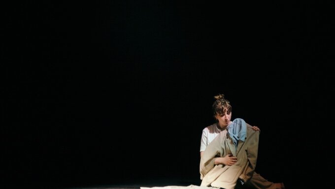 In a dark room a woman with a white t-shirt sits on the floor she is clutching a cream suit with a blue shirt underneath