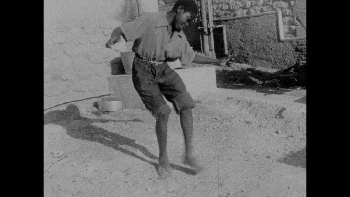A black and white photo of a young black man in a collared shirt with trousers rolled up to the knee, dancing barefoot.