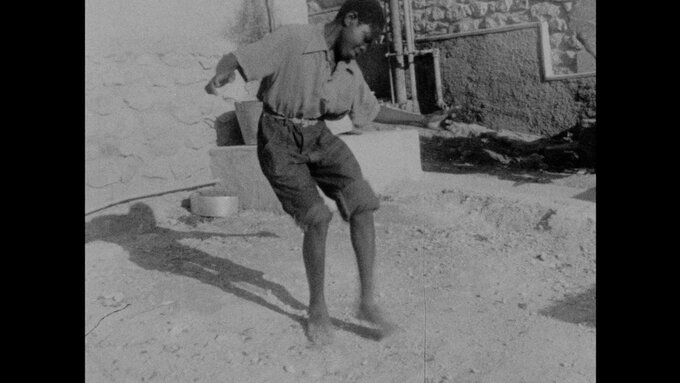 A black and white photo of a young black man in a collared shirt with trousers rolled up to the knee, dancing barefoot.