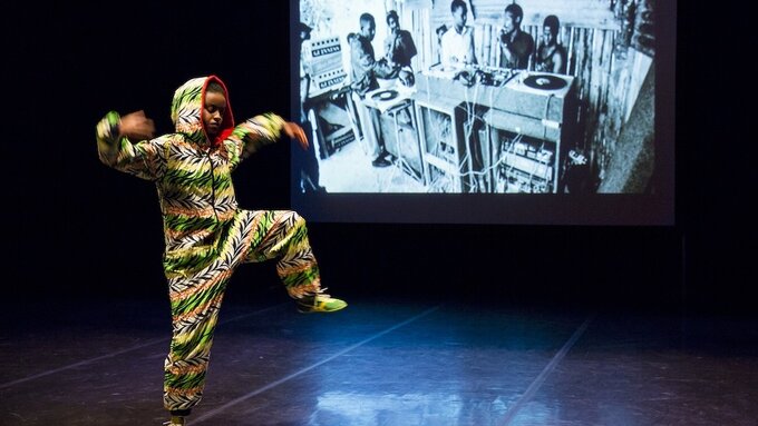 A performer dancing in front of a B&W image of people at turntable. They wear a hooded yellow & lime jumpsuit.