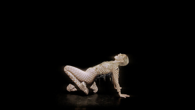 A performer crouches in an upward plank. Their hair is slicked back and wear a checker body suit with gold chainmail.