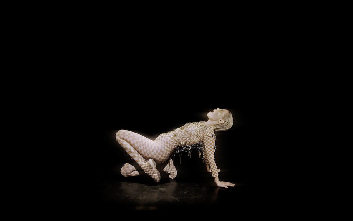 A performer crouches in an upward plank. Their hair is slicked back and wear a checker body suit with gold chainmail.