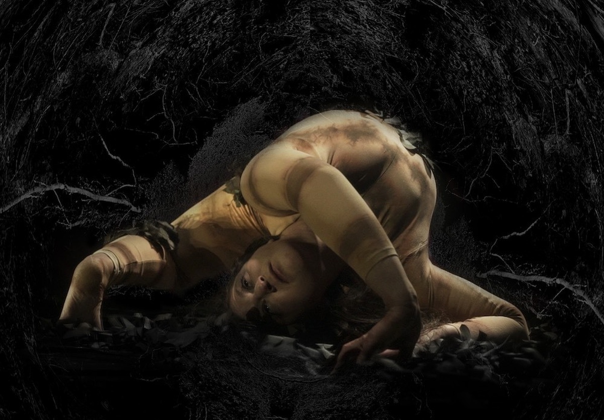 A performer contorts their body. In a flesh tone body suit. Behind them is a hole from which a matrix of plant roots.