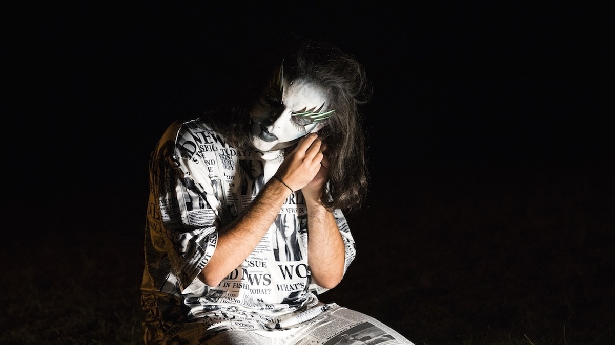 A drag performer with white face paint, a newspaper patterned suit, long green & black lashes crouched in a dark field.