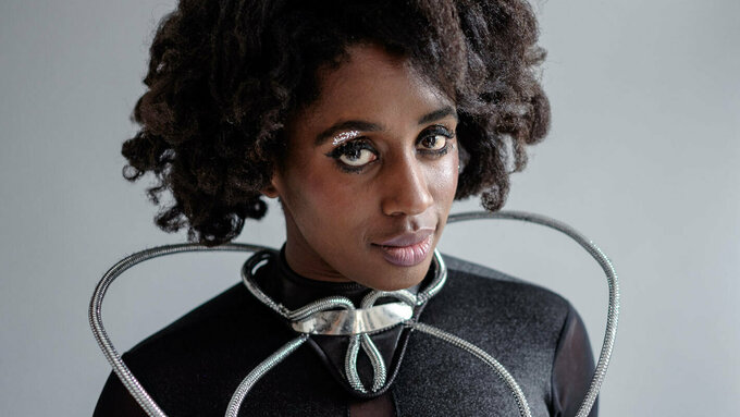 A an Afro-diasporic woman with silver eye shadow. She wears a sci-fi black top with silver tubes coiled around her torso