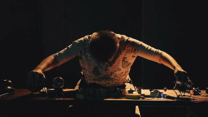 A person, head down, arms outstretched, head bent, is reaching out to the edges of a table laden with coils and springs.