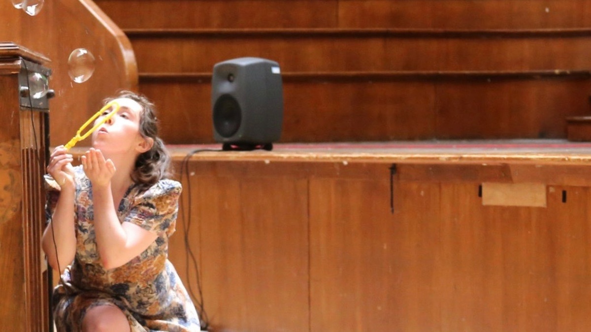 A woman in a flowery dress is kneeling down in a lecture hall, she is blowing bubbles through a plastic wand.