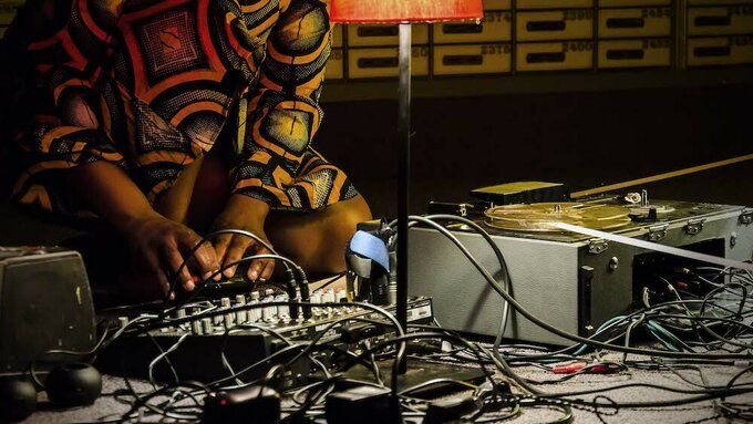The torso of a woman wearing a brightly patterned dress kneeling on a carpet with a mixer and a tape recorder.