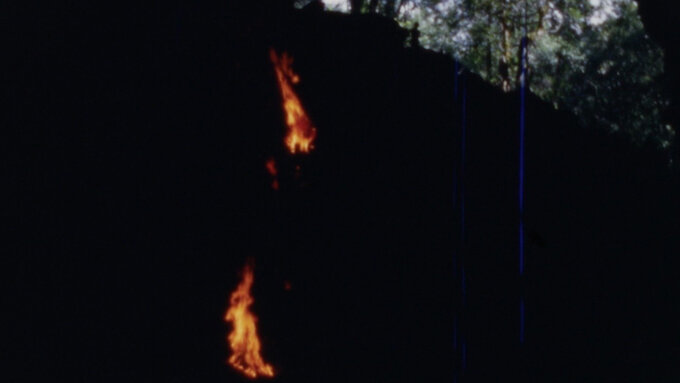 View from inside of a cave looking out to an opening where light can be seen. A number of flaming torches are visible.