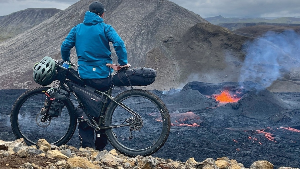 A photo of a man leaning against his bike, he's looking across a lava plane as lava and smoke emits from it