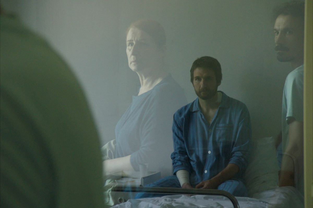 A man in a blue shirt is sat on a bed. In the foreground, we see reflections of woman and man look on through the glass.